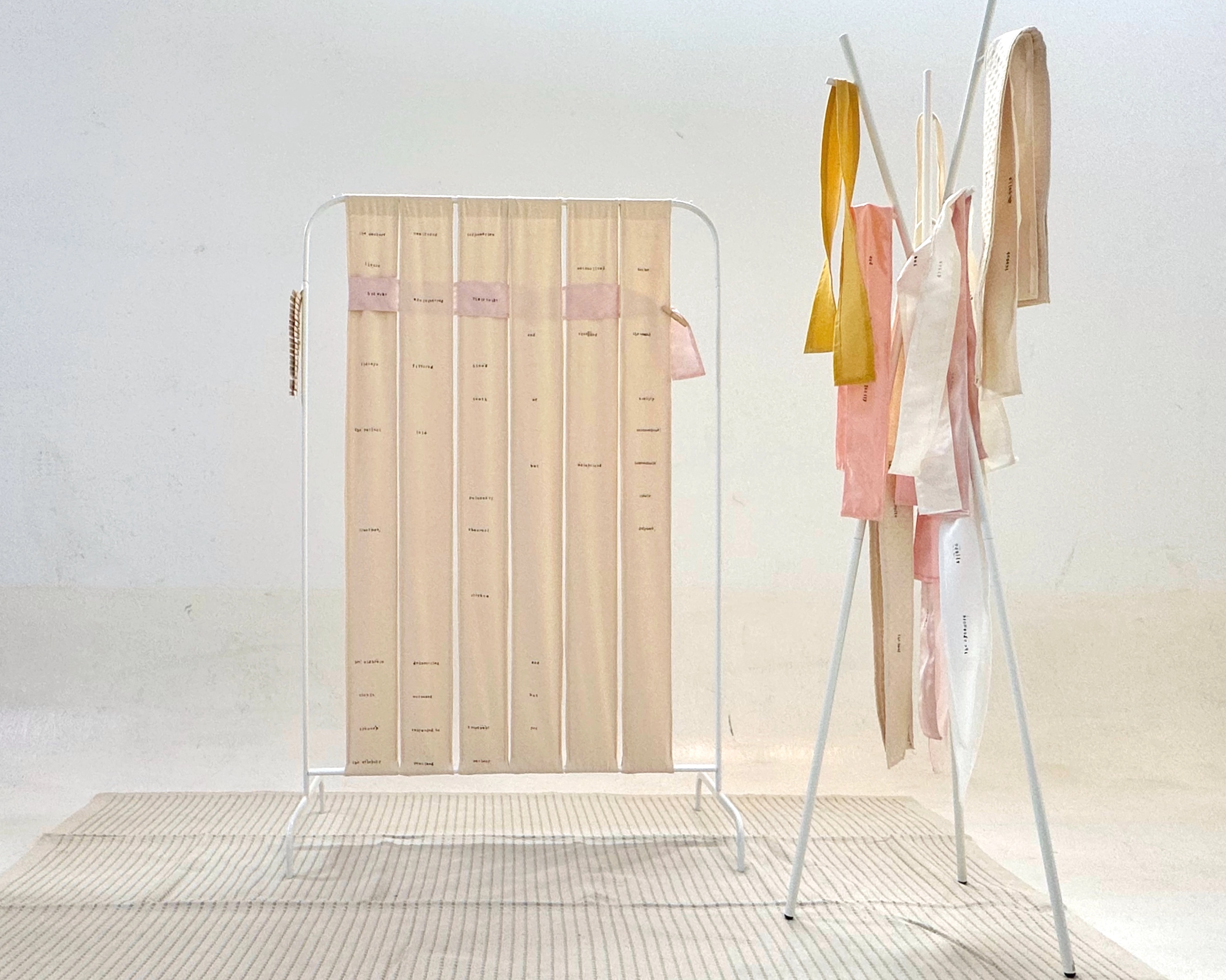 An installation featuring a woven textile piece displayed on a metal frame, with soft beige and pink tones, and strips of fabric hanging from a freestanding rack to the right.